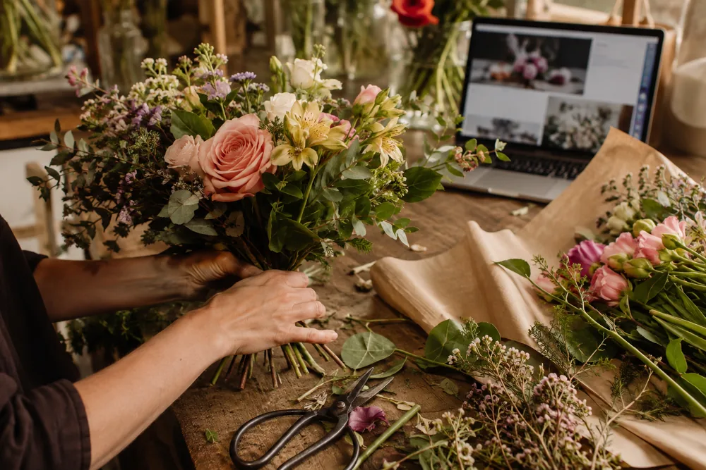 Manos montando un ramo en el taller de la floristería, con un portátil al fondo